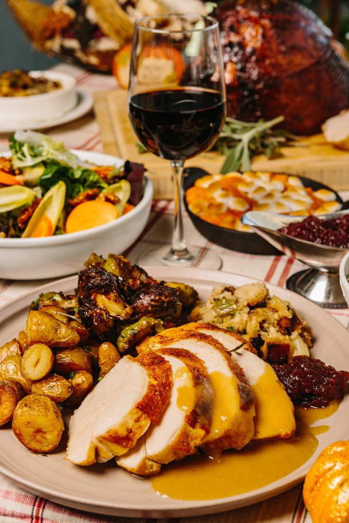 Side view of Thanksgiving feast plate complete with turkey and gravy, potatoes, brussels sprouts, stuffing and cranberry sauce. Glass of red wine and a fall table spread behind it.